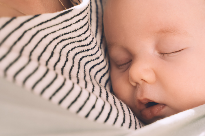 A close-up lifestyle shot of a sleeping newborn baby wearing a light sage green hooded wrap or carrier, nestled against a mother's striped black and white top.